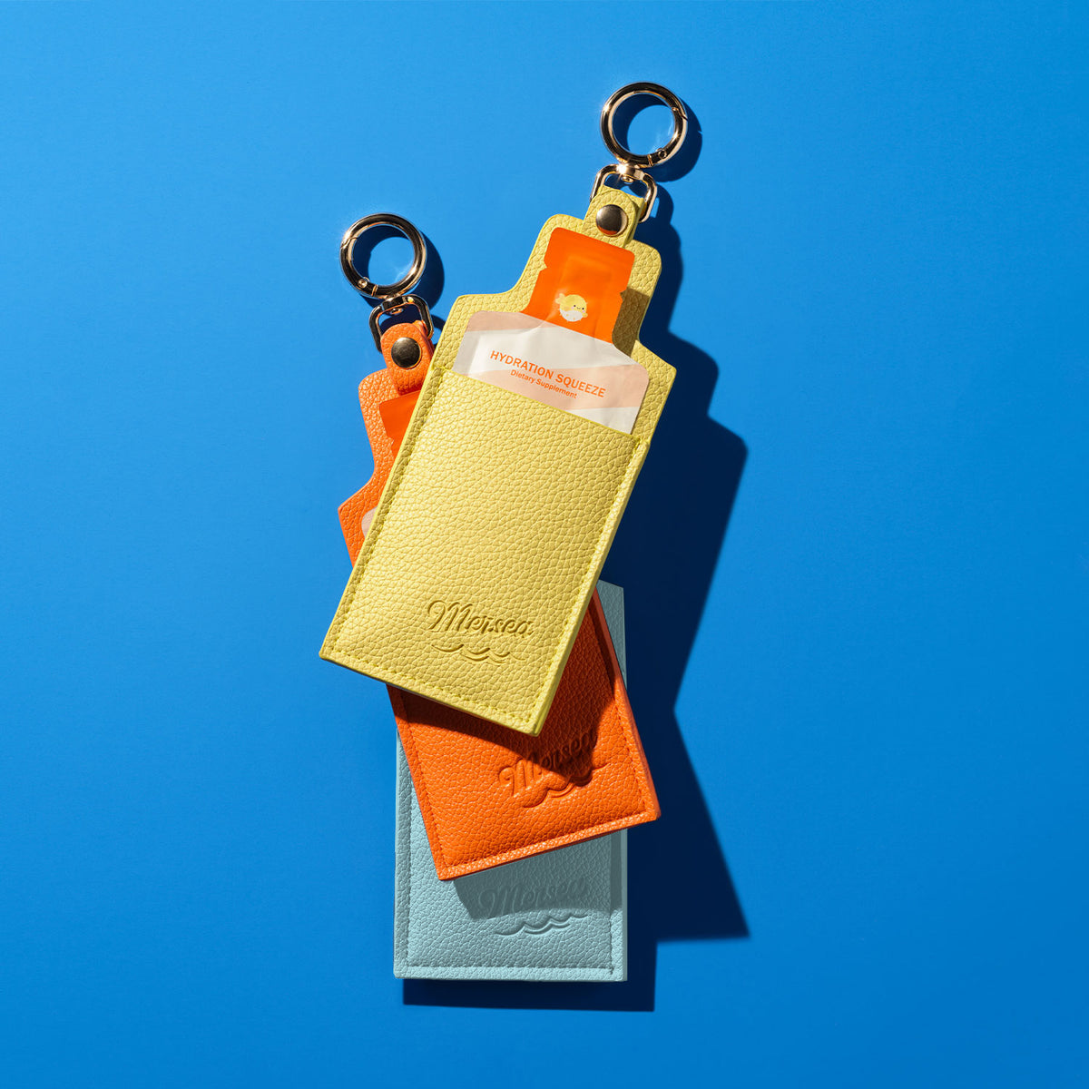 Stack of Mersea embossed keychain pouch holders in yellow, orange and blue with a Hydration Squeeze inside